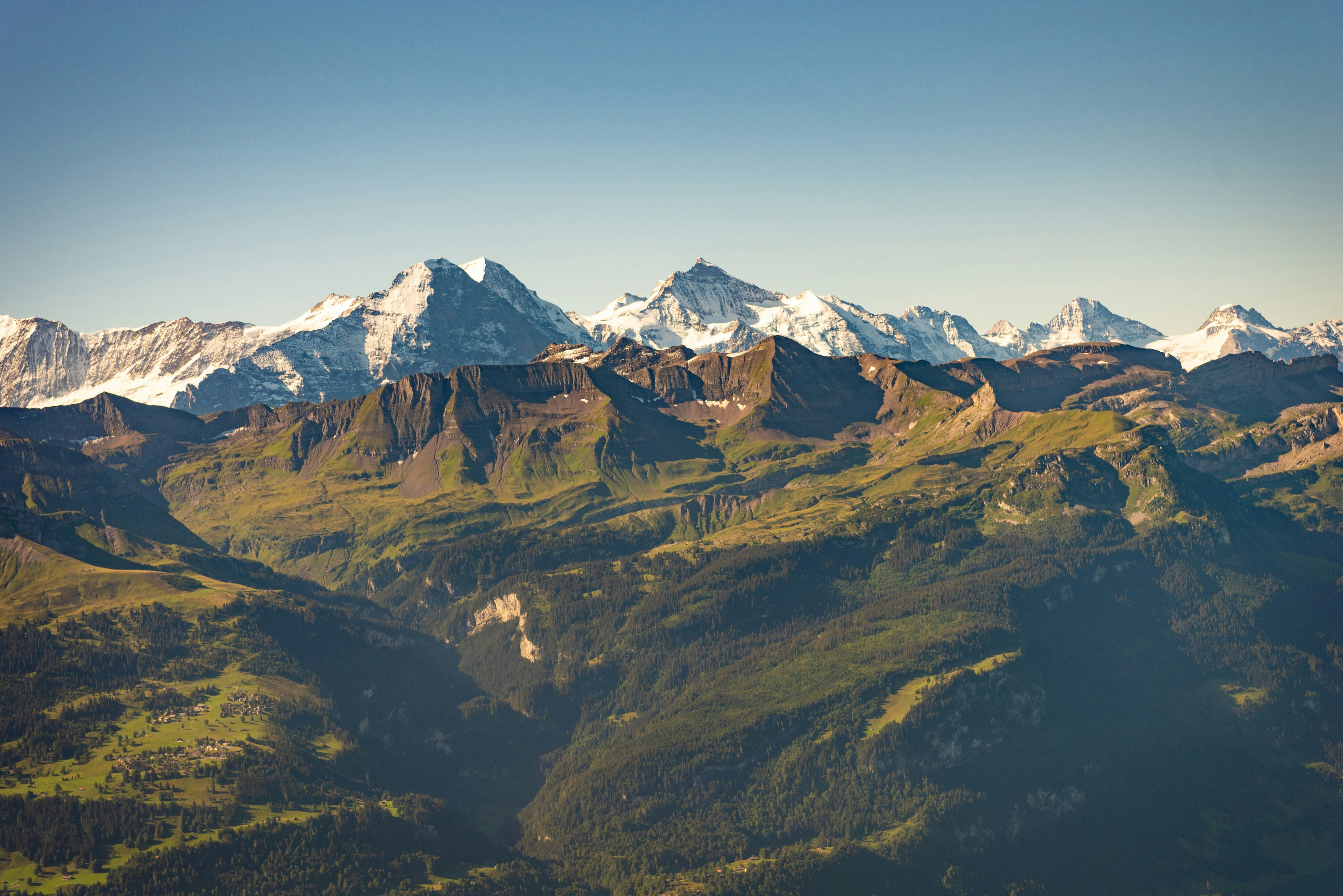 Mountain range with snow-capped peaks and green valleys under a clear blue sky.